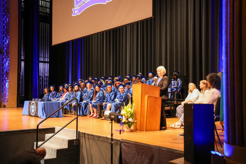 Principal Cindy McMahon speaks into a microphone on stage with graduates of The Community School.