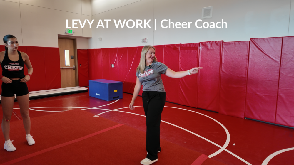Ferris Cheer Coach Emily Schutz smiles while directing student athletes in a gym with red mats.
