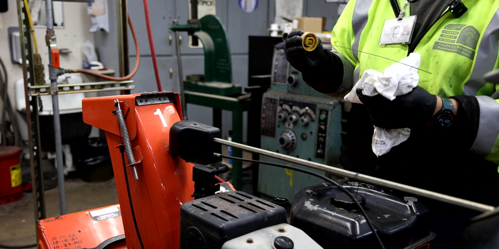 A maintenance worker in a high-visibility jacket, with gloves on, inspects and cleans a dipstick from a snow blower engine in a workshop setting.