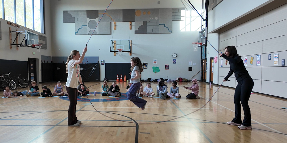 A student jumps rope with assistance from two adults in an elementary school gym.