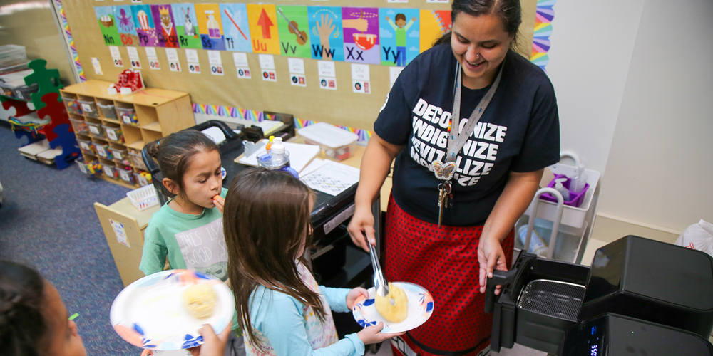 A teacher serves frybread to young students in a classroom. She is wearing a black t-shirt with the words "Decolonize, Indigenize, Revitalize" printed on it and a red ribbon skirt.