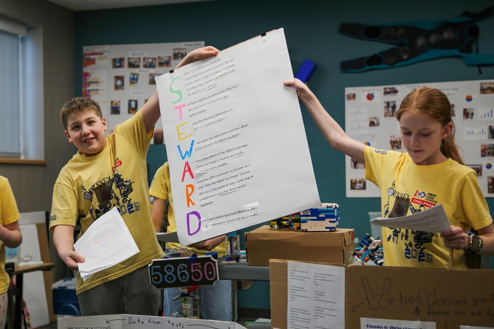 Boy and girl hold up poster during presentation. Poster reads "STEWARD" in large font beside smaller, unreadable font.
