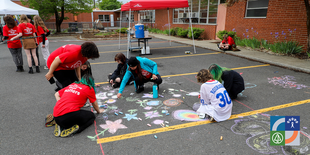High school students at the community school wearing red shirts with the text “Do Something That Matters” on the back create chalk art in a parking lot; tents are set up around them as part of an outdoor festival.