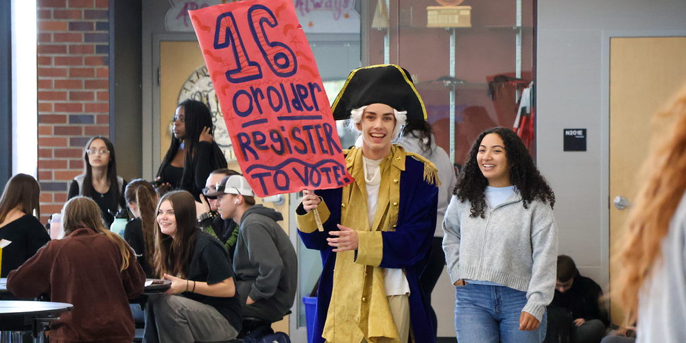 North Central students, one dressed as George Washington, carry a sign reading “16 or older, Register to vote” in a high school cafeteria.