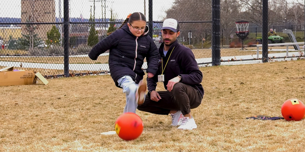 An adult helps a visually impaired student kick a red ballon a school field.