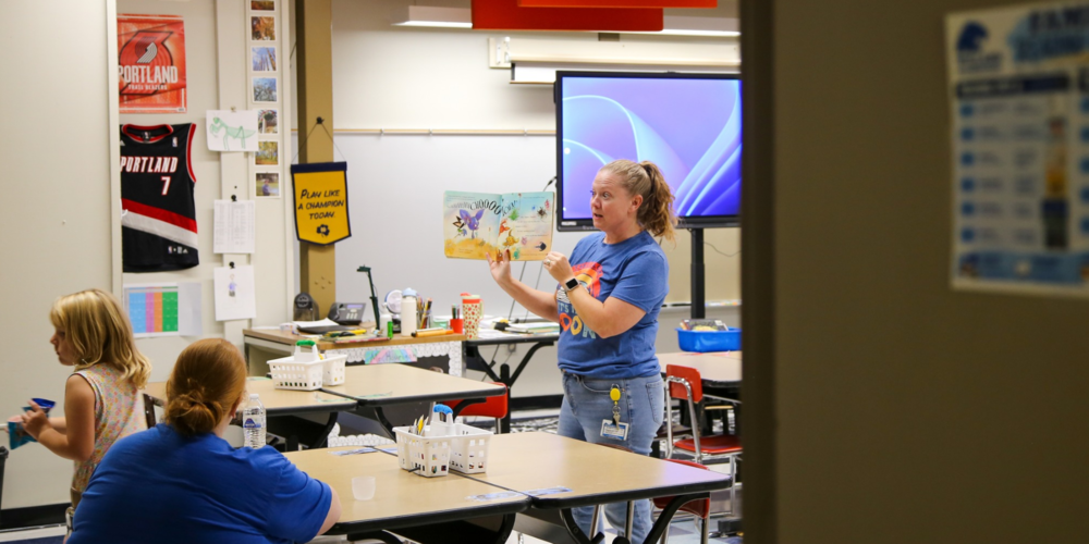 An educator stands at the front of an elementary school classroom holding open a book while reading to a group of children and adults.