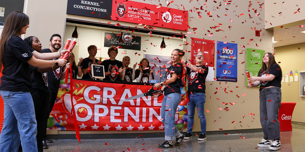 A group of people stand in front of a counter decorated with a large red banner that reads “Grand Opening” in bold white letters. The counter has signage for North Central High School and Gesa Credit Union. Red confetti fills the air as two individuals hold oversized scissors and cut a red ribbon, while others hold confetti poppers.