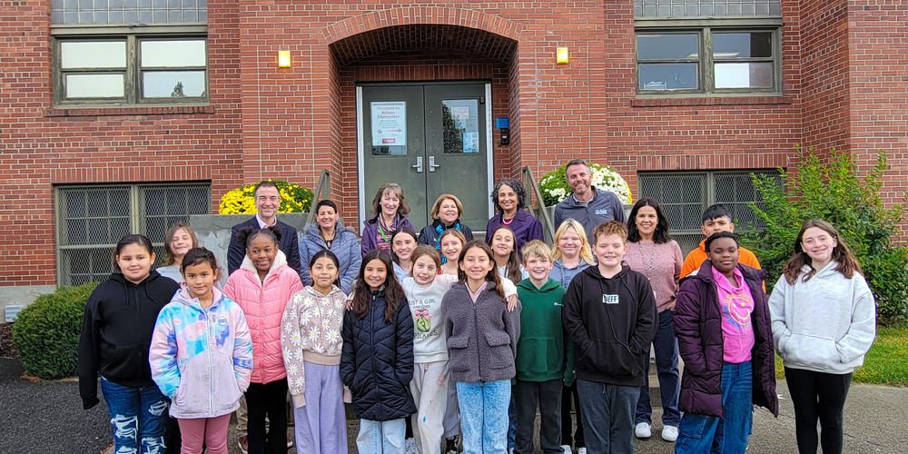 A large group of fifth grade students stand with city and school leaders outside a brick building.