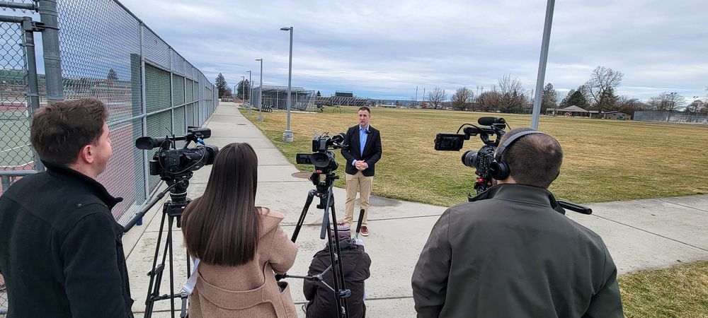 Man standing in front of three news cameras, with field in background.