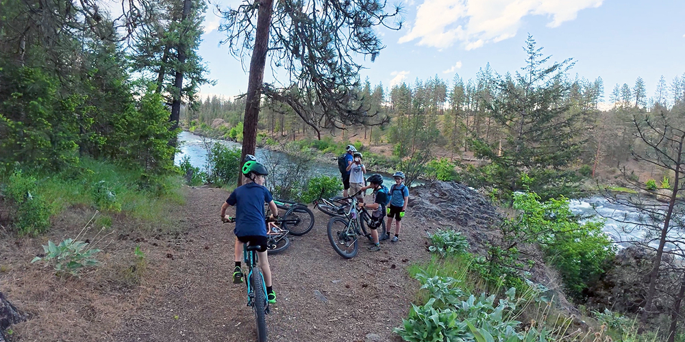 Children riding bikes stop at an outlook next to a river in a wooded area.