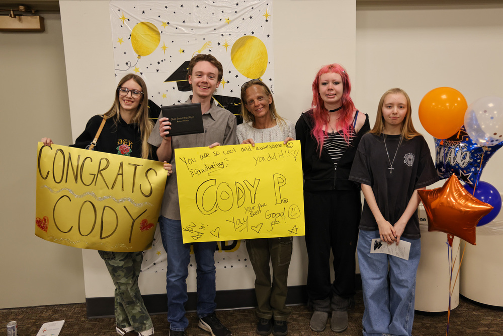 A Project SEARCH graduate holds their diploma with family members who are holding Congrats, Cody signs and balloons.