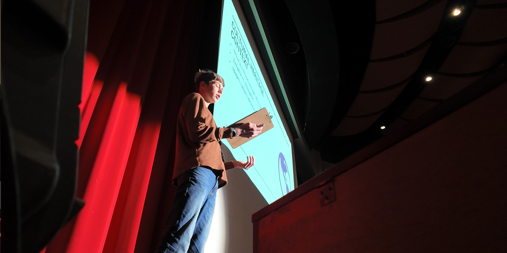 A student stands in a spotlight on stage for a presentation in a high school auditorium.