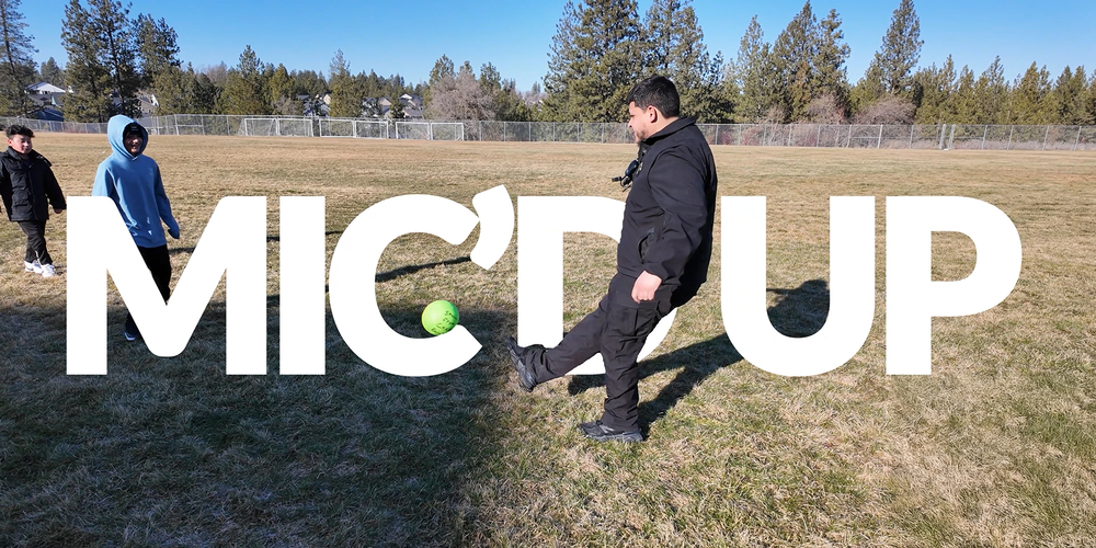 A Campus Safety Specialist kicks a soccer ball to Chase Middle School students on a grass field. Accompanying text is Mic’D Up
