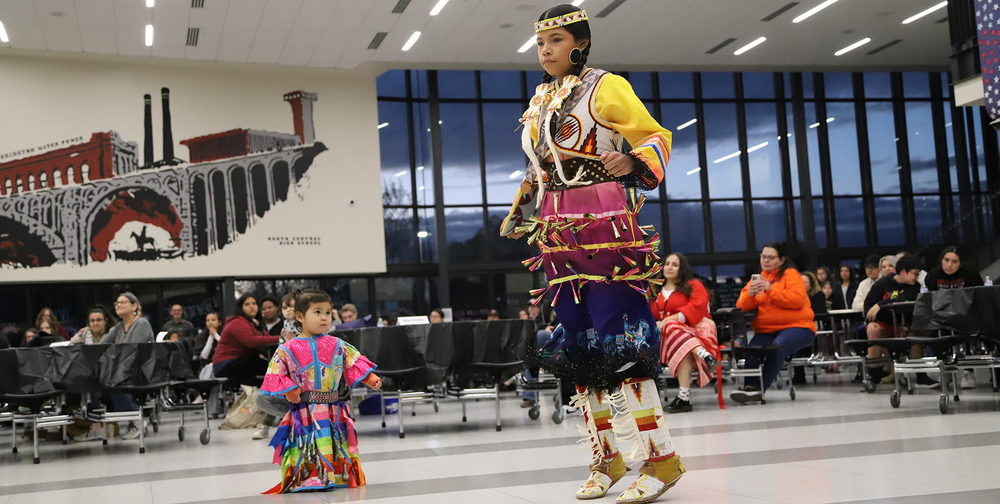 Two Native American girls, a middle school student and a preschooler dressed in jingle dresses and regalia, dance in a high school commons. Audience members watch at tables behind them. A mural of central Spokane are on the wall behind them, as are windows turning dark with sunset.