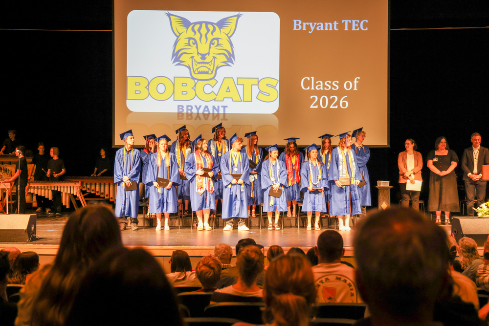 Graduates in blue gowns and caps stand on a stage in front of a screen displaying the Bryant/TEC Bobcats logo and "Class of 2026".
