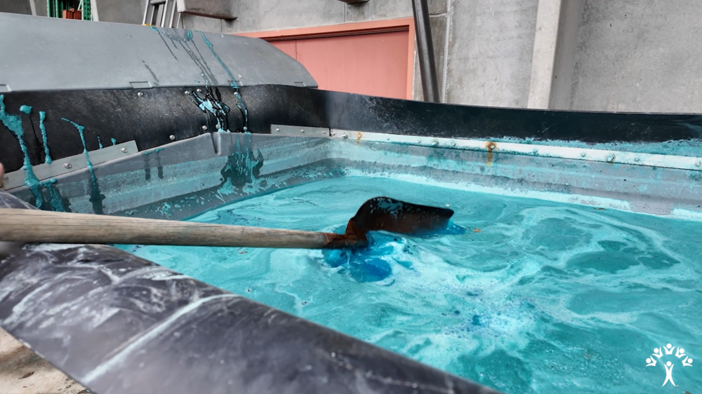 A shovel stirs a blue liquid in a large, open vat.