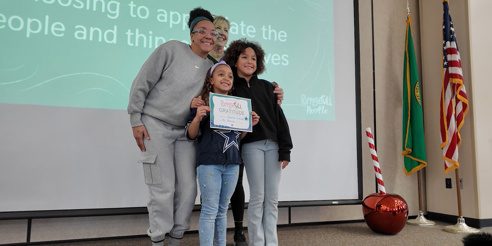 A student smiles with an award with two adults and a peer on stage at an elementary school.