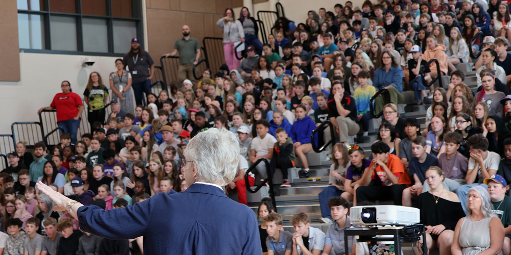 A woman with white hair speaks to bleachers full of middle school students.