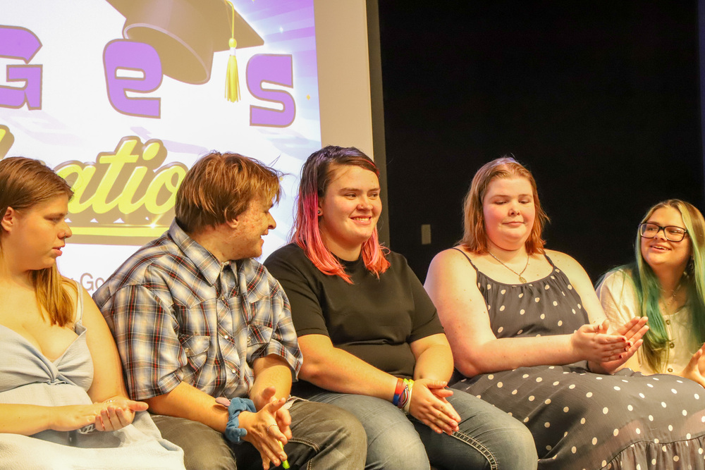 A group of young people sit on a stage in front of a screen with graduation text and a graduation cap graphic. They are smiling and clapping.