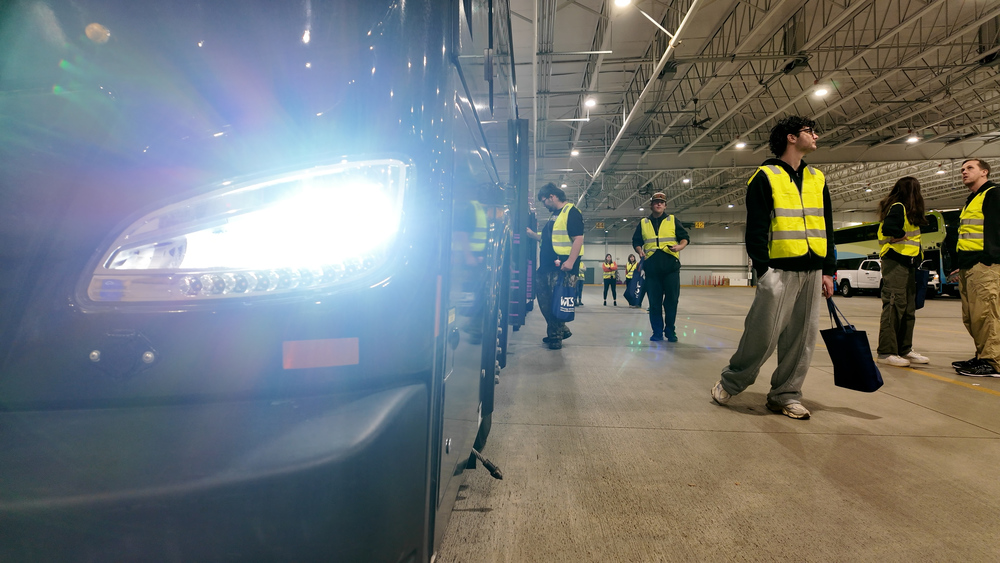 Students wearing yellow vests look around a bus garage and a city bus with its headlights on.