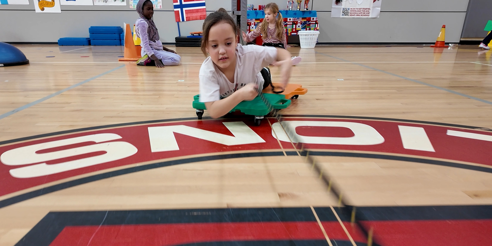 A Linwood Elementary School student pulls on a rope and slides across the gym floor on a scooter during a P.E. activity.
