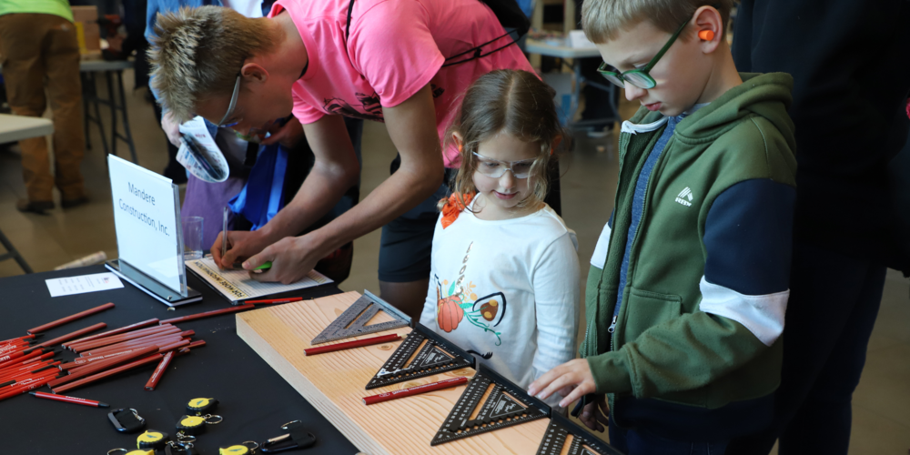 Two children wearing orange ear plugs and safety googles look and touch a set square on a wood block at a trade show; an older student in a neon pink shirt fills out a contact sheet next to them.