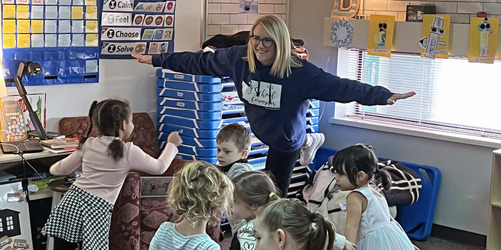 A school counselor and a group of elementary students stands on one leg and holds their arms out like they are flying in a classroom activity.