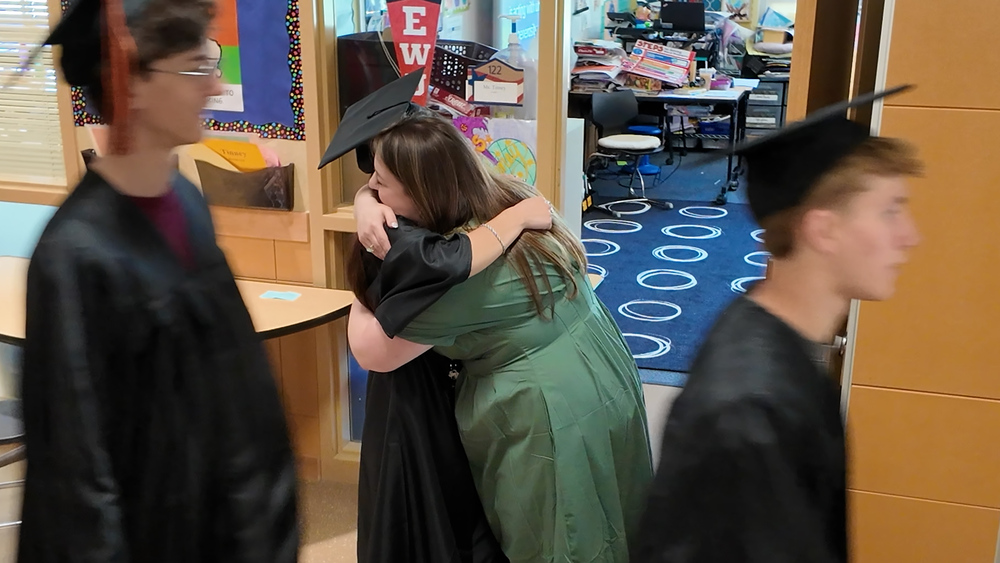 An elementary teacher hugs a high school senior in a graduation cap and gown in an elementary school hallway.