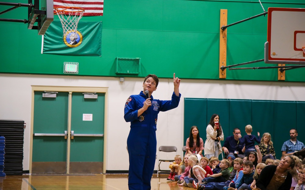 Astronaut Anne McClain points toward a projector screen in an elementary gymnasium. American flag in background.