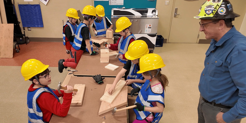 Mullan Road elementary students in hard hats and blue vests hammer nails into wood while an adult volunteer supervises.