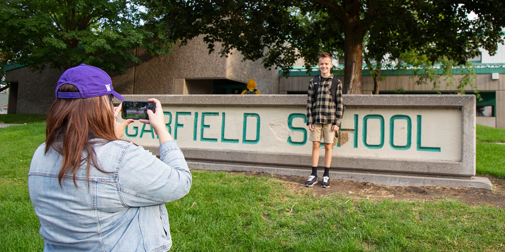 A parent takes a photo of a student in front of a school sign.