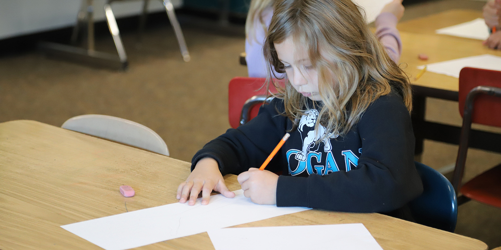 An elementary student draws on a paper with a pencil in a classroom.