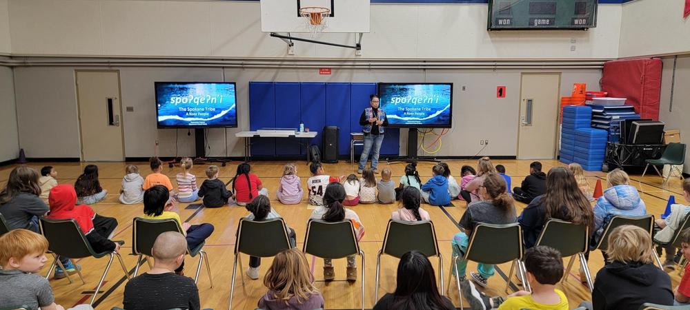A member of the Spokane Tribe of Indians speaks to a group of students seated in the gymnasium at Regal Elementary.