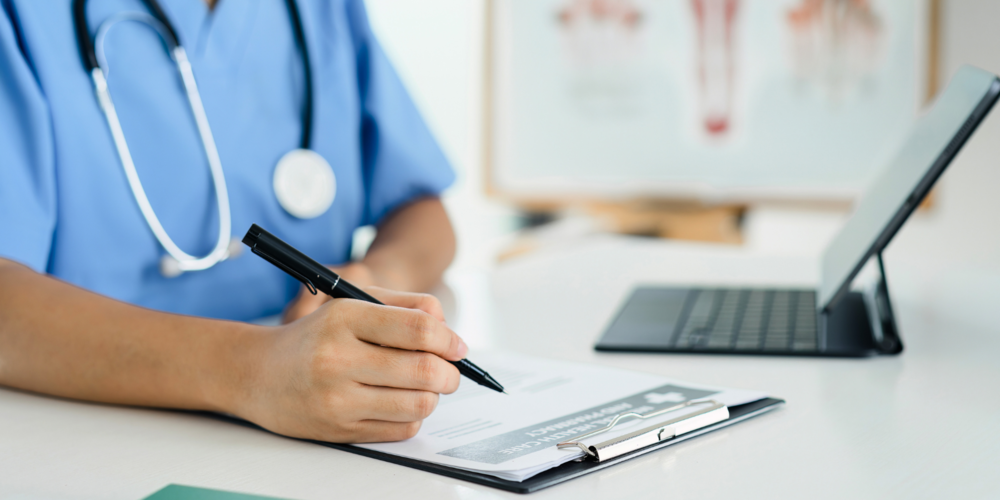A medical professional makes notes on a clipboard next to a laptop.