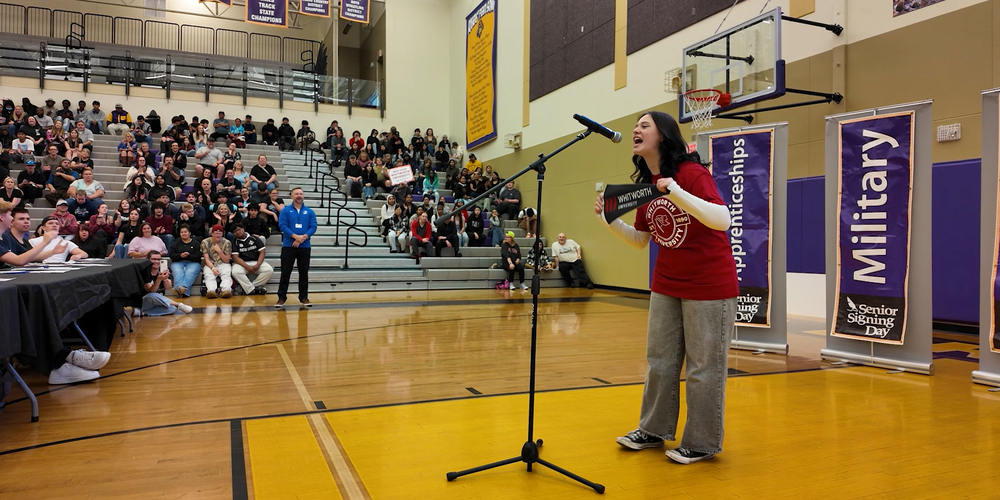A Rogers High School student wearing a Whitworth University t-shirt and holding a pennant speaks into a microphone with gym bleached filled with audience members.