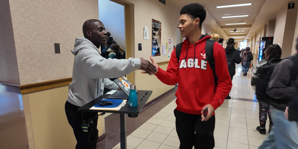 A smiling principal greets a student in the hallways during a passing period from a mobile desk station with a laptop, computer mouse, and water bottle.