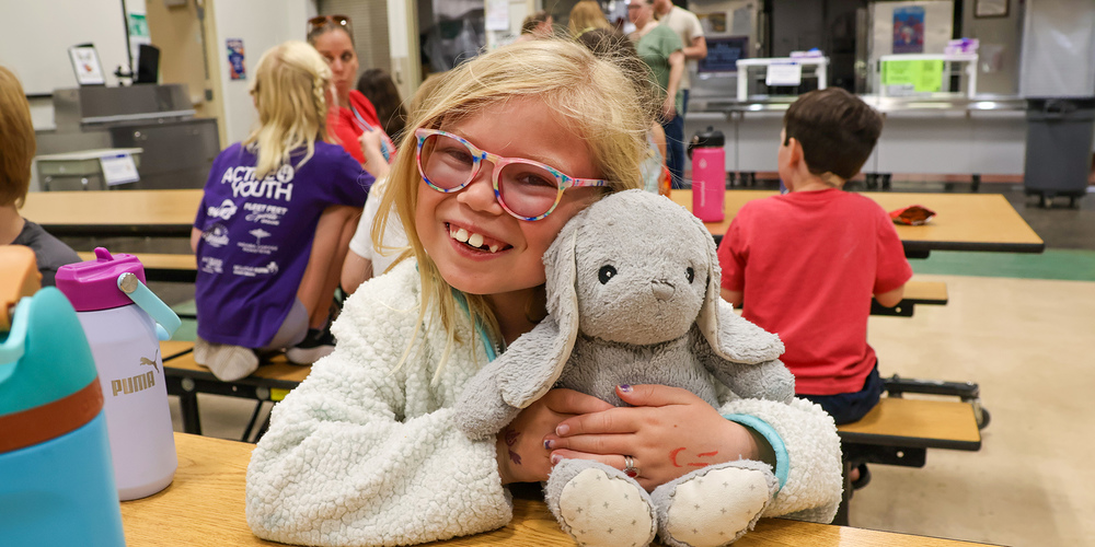 An elementary student smiles and cuddles a plush toy bunny in a cafeteria.