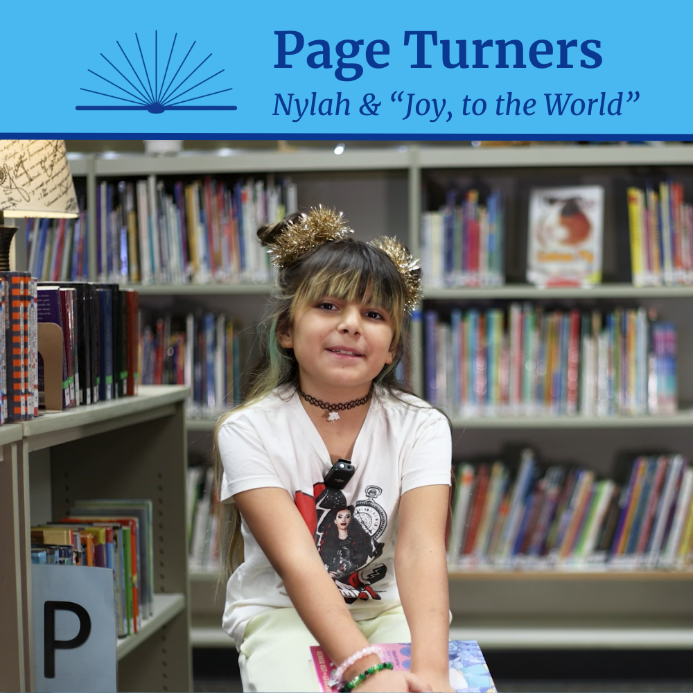 A girl holding a book in an elementary school library with the accompanying text “Page Turners: Nylah & ‘Joy, to the World’”