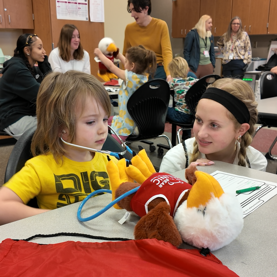 Preschool student uses a stethescope to listen to a plush eagle's heartbeat with a high school student.