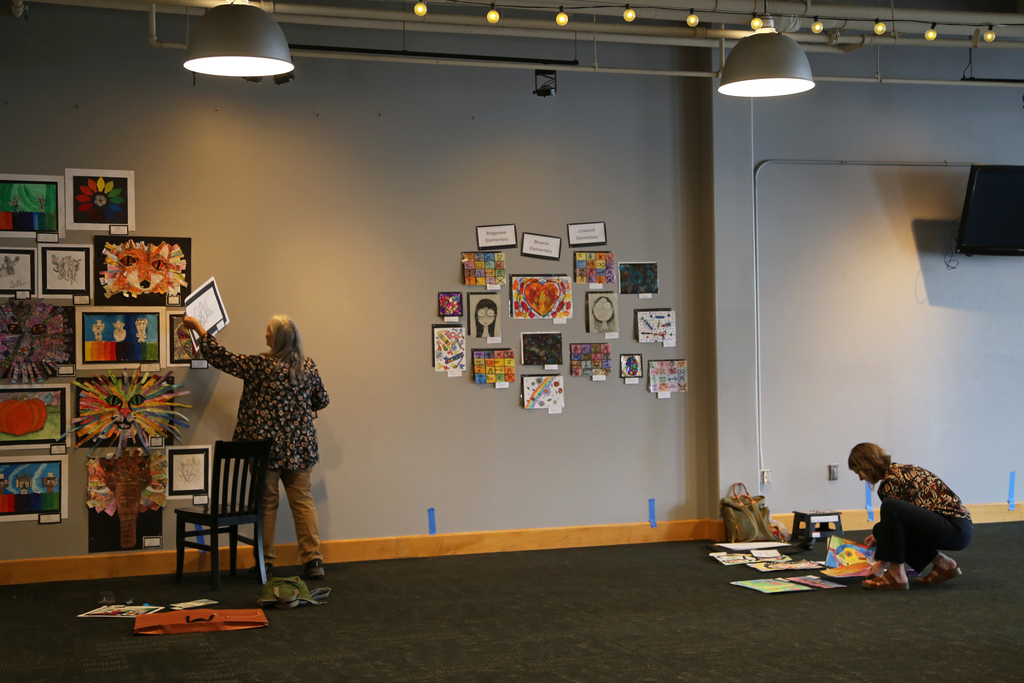 Two teachers hang student artwork in a gallery display.