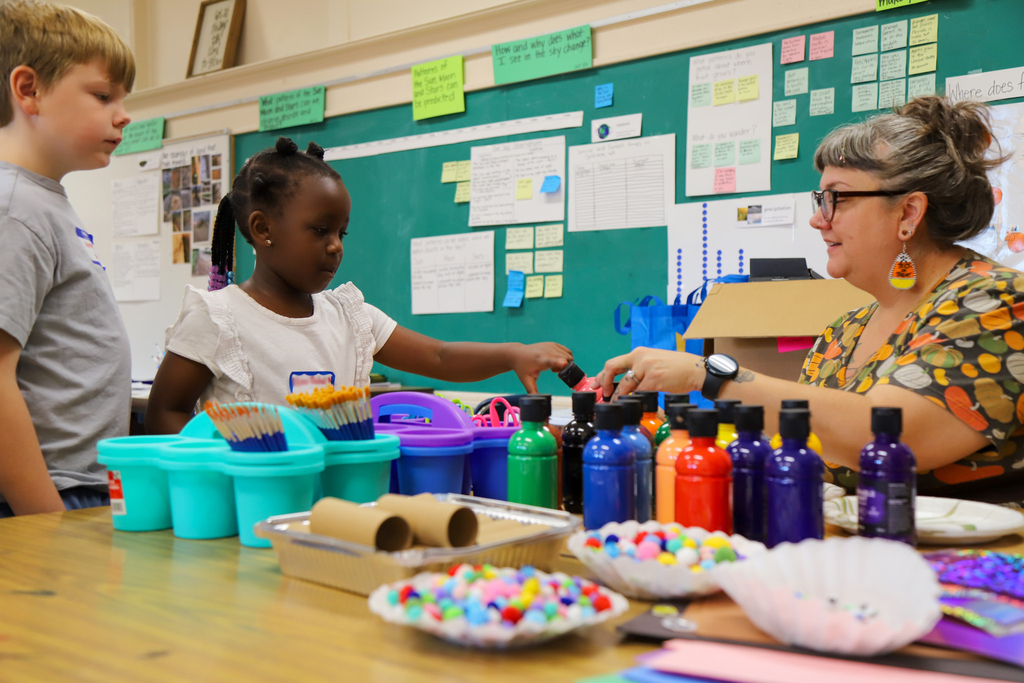 An adult helps elementary students with art supplies in a classroom.