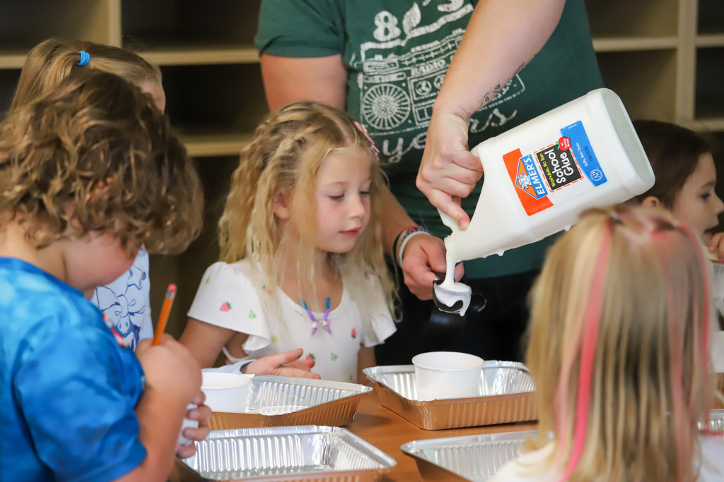 An adult pours glue for students who are making slime in a classroom setting.