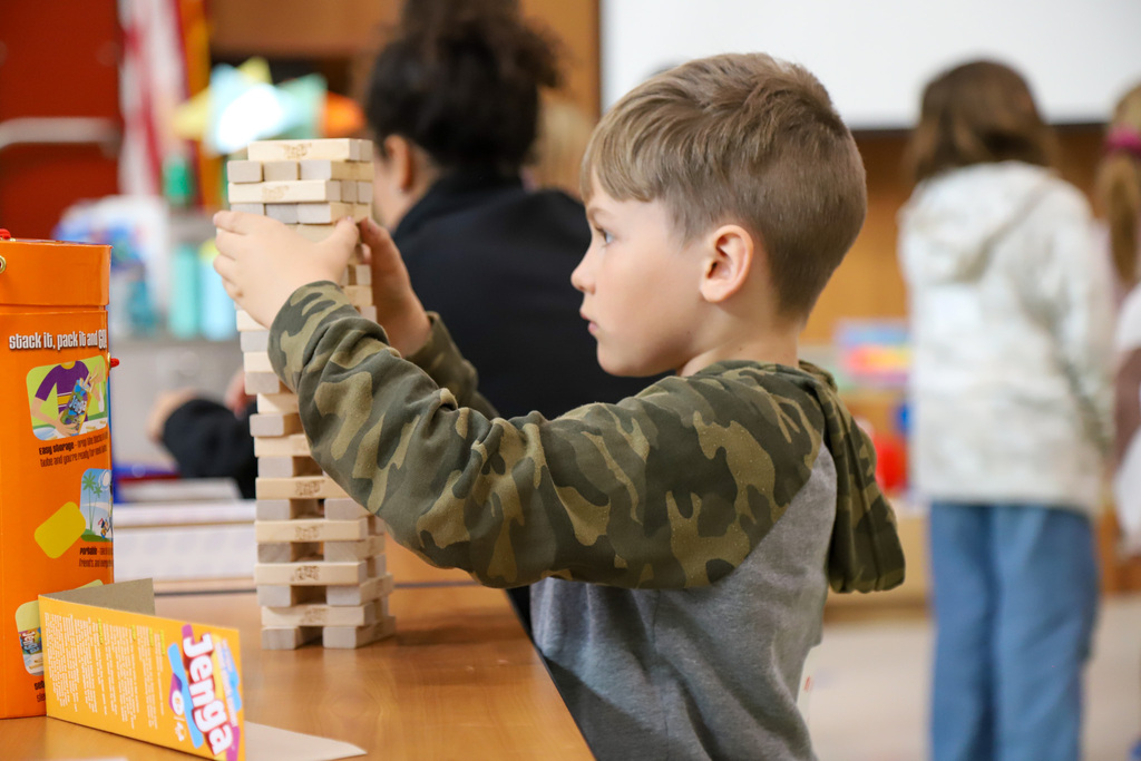 An elementary student builds a Jenga tower in a room with other students.