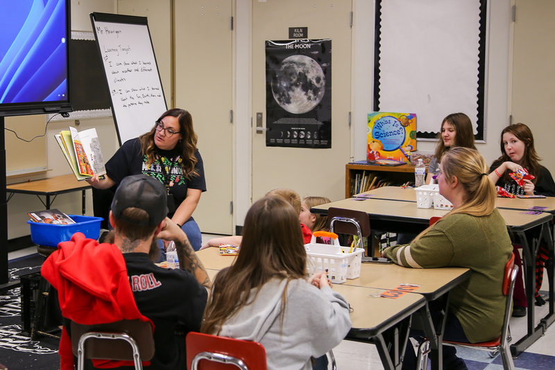 An educator stands at the front of an elementary school classroom holding open a book while reading to a group of children and adults.