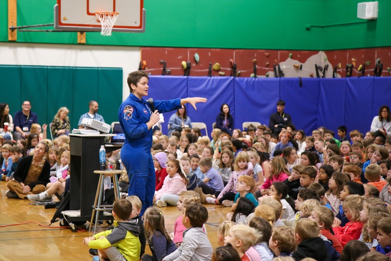Astronaut Anne McClain gestures to group of elementary students in school gym.