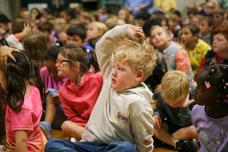 Young boy with blonde hair raises hand.