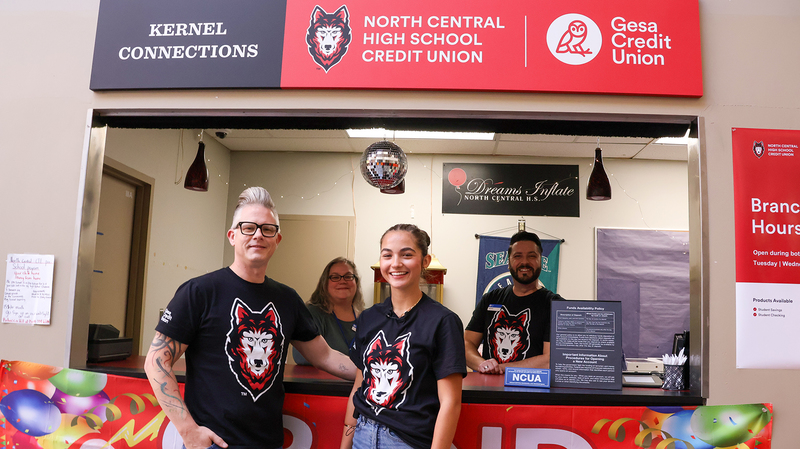 Two people stand in front of a counter with a “North Central High School Credit Union” sign above. The counter displays informational materials and decorations, including a disco ball overhead and colorful balloons on the banner.