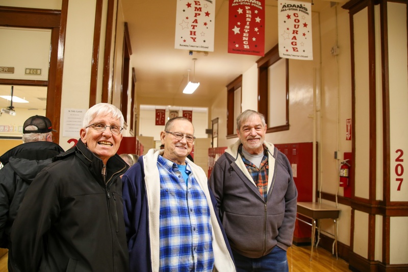 Three older men stand in the hallway of a school, smiling at the camera.