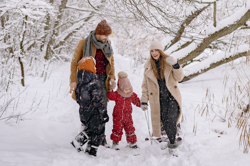 A family of four in winter coats, scarves and hats walks and laughs on a snowy day in a wooded area.