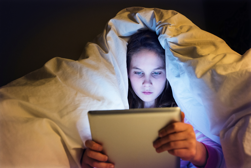 A girl hides under a blanket at night, illuminated by the glow of a tablet screen in her hands.